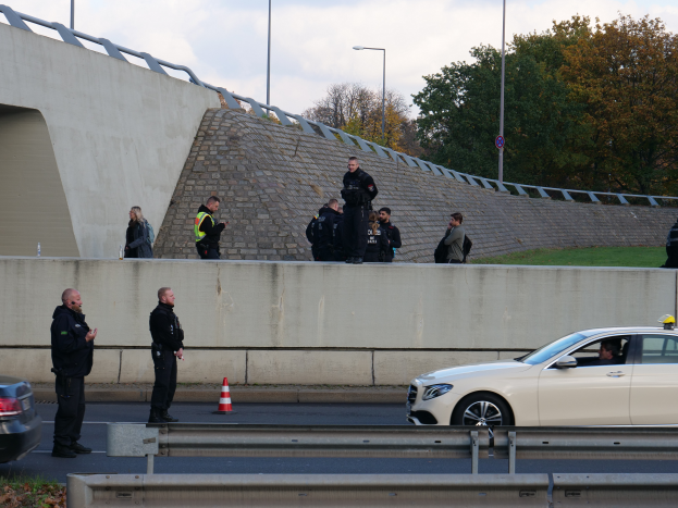 Eine Gruppe von Polizeibeamten steht an der Straßenseite neben einem Auto, mit Verkehrskegeln, einer Trennwand, Gras, einer Wand, Laternenmästen, Bäumen und einem bewölktem Himmel im Hintergrund.