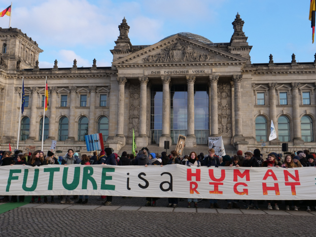 Eine Gruppe von Menschen hält ein Transparent mit der Aufschrift "Zukunft ist Menschenrecht" vor dem Reichstaggebäude in Berlin, Deutschland, mit sichtbaren Fenstern, Säulen und Bögen des Gebäudes, umgeben von Fahnenmasten und einem bewölkten Himmel.