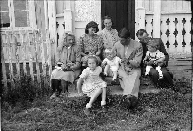 Ein Schwarz-Weiß-Foto einer Familie, die auf den Stufen eines Hauses sitzt und steht, wobei eine Person ein Baby hält, ein Tür und ein Fenster im Hintergrund und Gras auf dem Boden