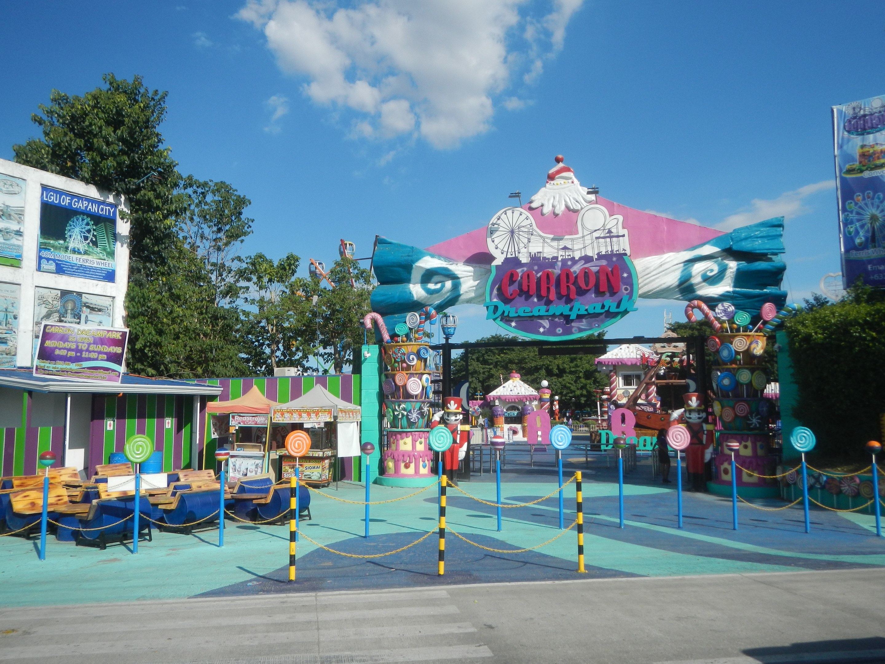 Kirmesattraktion am Parkeingang mit Straße, eingezäuntem Bereich, Buden, Schildern, Bäumen und bewölktem Himmel im Hintergrund.