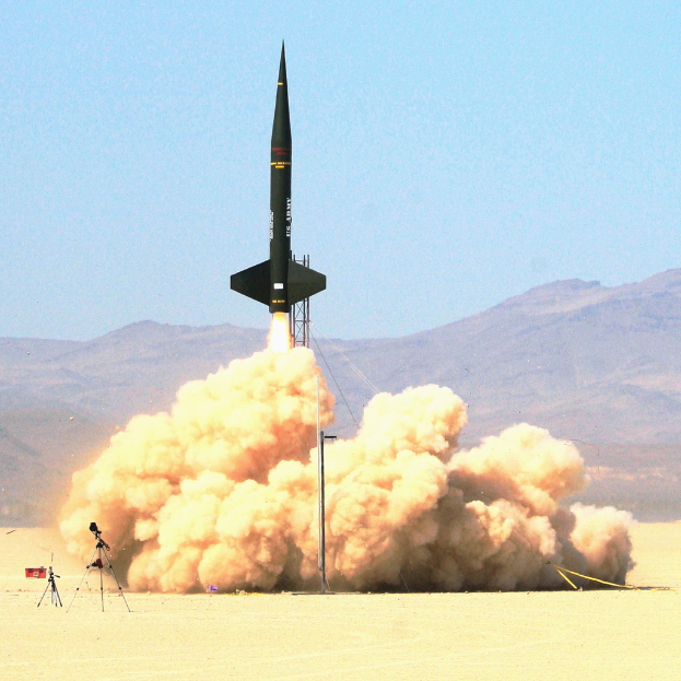 Eine Rakete startet vom Boden aus, steigt in einen klaren Himmel auf, mit einer Rauchspur im Schlepptau, vor einer Kulisse von fernen Bergen.