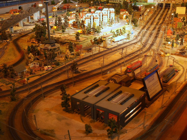Eine detaillierte Miniatur-Modellbahnausstellung in einem Museum, mit Gebäuden, Bäumen, Eisenbahnschienen, Fahrzeugen, Polen, Lampen und anderen Gegenständen, die eine belebte Stadtlandschaft darstellen.