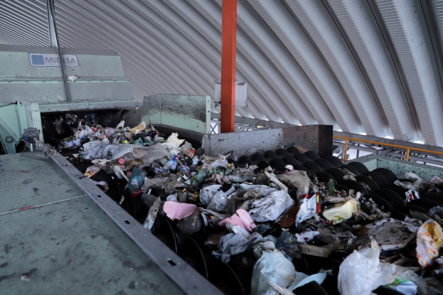 Großer Müllberg auf einem Förderband in einer Recyclinganlage mit verstreutem Müll und einem Metalldach oben.