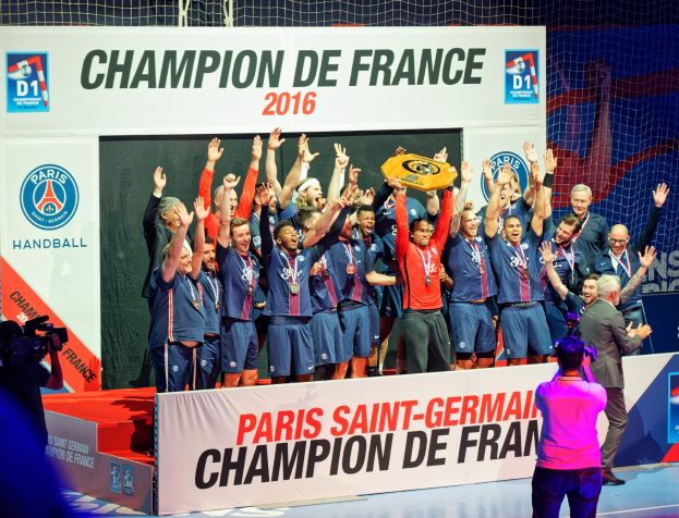 Eine Gruppe von Männern auf einer Bühne mit einem Pokal, einer Person, die filmt, und einem 'Champion de France 2016'-Plakat im Hintergrund.