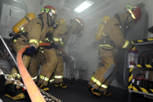 Feuerwehrleute in Schutzausrüstung mit Helmen und Sauerstoffflaschen arbeiten an einem Löschfahrzeug, mit einem Gestell voller Gegenstände und industriellem Hintergrund.