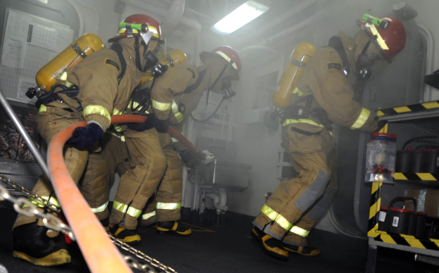 Feuerwehrleute in Schutzausrüstung mit Helmen und Sauerstoffflaschen arbeiten an einem Löschfahrzeug, mit einem Gestell voller Gegenstände und industriellem Hintergrund.