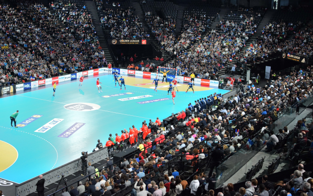 Eine Gruppe von Menschen spielt Handball in einer großen Arena bei Nacht, mit Zuschauern auf den Tribünen und einem Netz, das das Spielfeld teilt.
