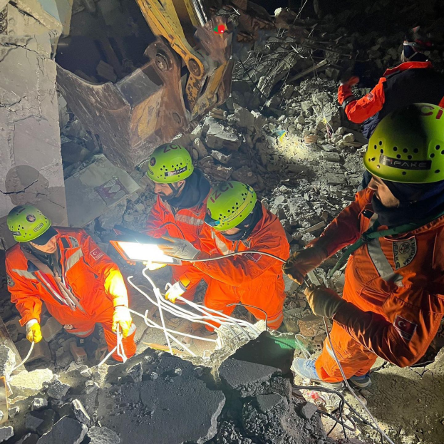 Eine Gruppe von Rettungskräften in orangefarbenen Uniformen und grünen Helmen beseitigt Trümmmer von einem eingestürzten Gebäude mit einem Kran im Hintergrund, höldend Rettungsgeräte und Materialien.