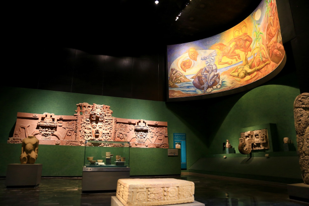 Ein Museumsausstellungsraum mit verschiedenen Artefakten, darunter Skulpturen, Tischgegenstände, eine Texttafel, eine Wandmalerei und Deckenleuchten im Mexiko-Stadt-Museum der Anthropologie.