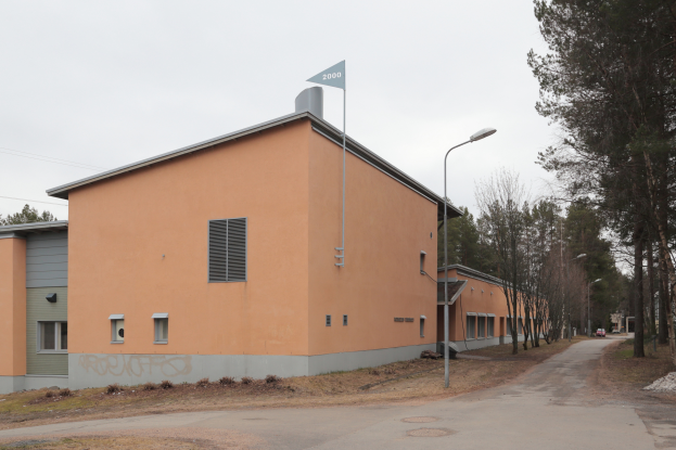 Ein orangefarbenes Gebäude mit einer Fahne oben drauf, umgeben von Straßenmasten, Straßenlaternen, Kraftfahrzeugen, Bäumen und einem klaren blauen Himmel.