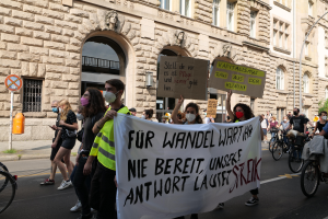 Eine Gruppe von Menschen marschiert auf einer Straße in Berlin, hält Schilder und Banner hoch und fährt mit Fahrrädern, vor einem Gebäude mit Fenstern, Bögen, Säulen, Skulpturen, Bäumen und einem Laternenmast, bei einer Demonstration gegen die Anti-Kriegsbewegung.
