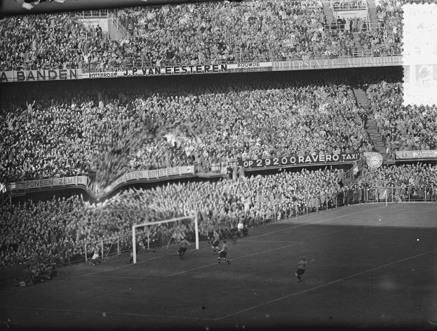 Ein Schwarz-Weiß-Foto eines Fußballspiels in einem Stadion, mit Spielern auf dem Boden, einem Torpfosten, einem Zaun, einer Fahne mit Text, einer Treppe und einem Dach mit Deckenbeleuchtung.