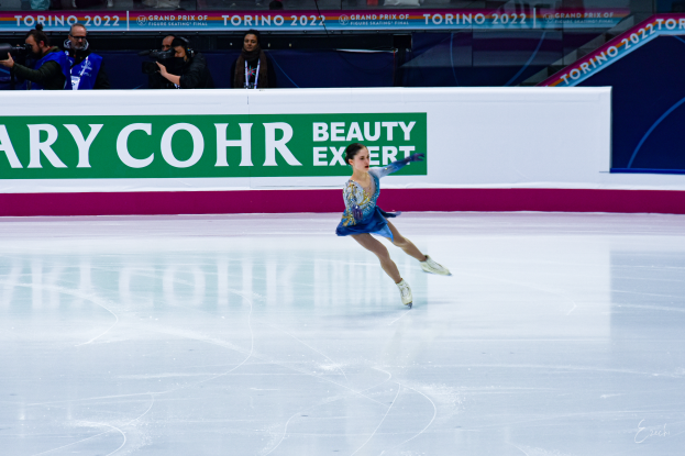 Eine Frau in einem blauen Kleid Schlittschuh auf einem Eisstadion, umgeben von Zuschauern mit Kameras, mit einem Schild im Hintergrund, auf dem "Tessa Virtue und Scott Moir" steht.