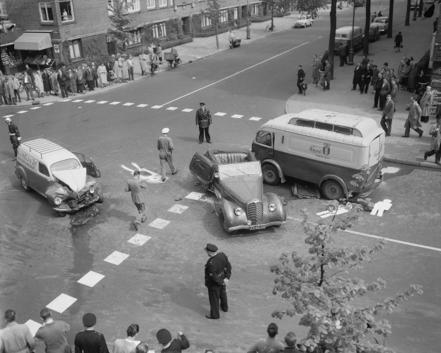 Schwarz-weiß-Szene eines Autounfalls an einer Straßenkreuzung mit mehreren Fahrzeugen, darunter ein Van, und einer Gruppe von Schaulustigen; Gebäude, Bäume, Pfähle und Texttafeln im Hintergrund.