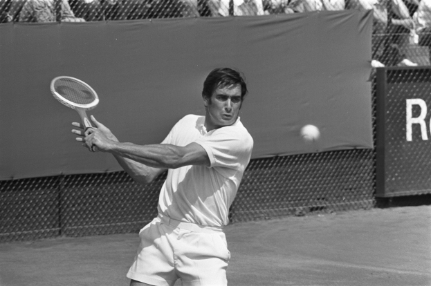 Ein Mann schlägt mit einem Tennisracket nach einem Ball während eines Matches, mit Zuschauern auf den Rängen und einem Banner im Hintergrund.