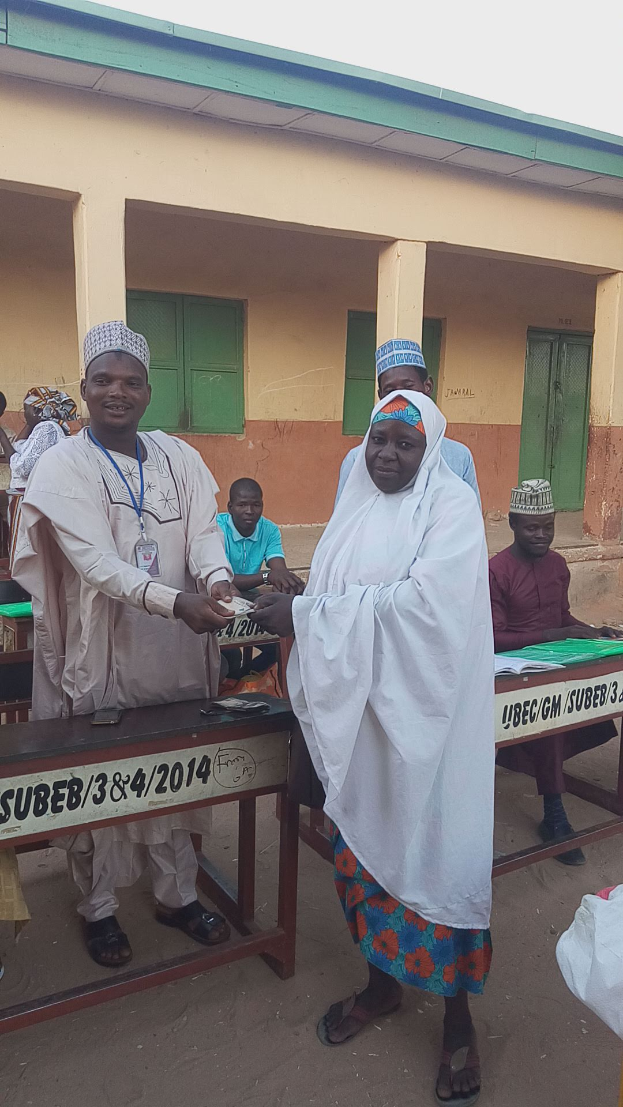 Eine Gruppe von Menschen steht und sitzt um einen Tisch mit Papieren und Gegenständen herum, vor einem Gebäude unter einem klaren blauen Himmel, die scheinbar bei einer Wahlurne wählen.
