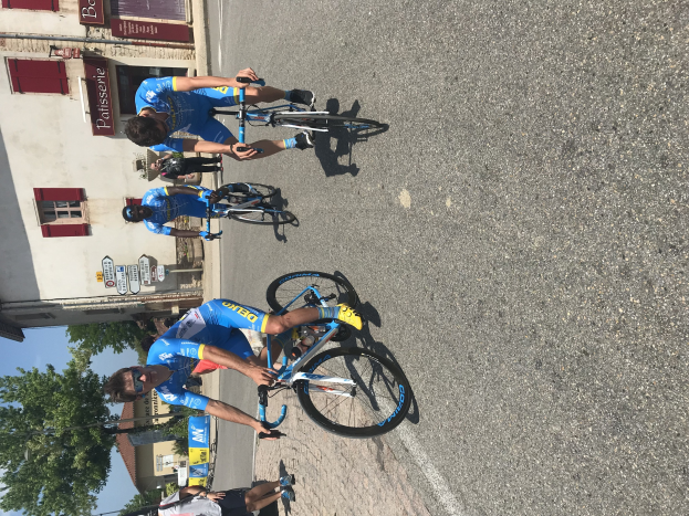 Eine Gruppe von Radfahrern fährt eine Straße entlang an einem Gebäude mit Fenstern und Schildern vorbei, mit Bäumen und einem blauen Himmel im Hintergrund und Zuschauern auf der Straße darunter.