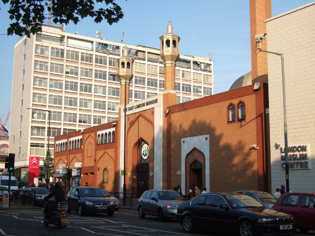 Ein Moscheebau mit Fenstern und Glastüren, mit einem Namensschild an der Wand, Straßenlaternen, einer Ampel an einem Mast, Fahrzeugen auf der Straße, Gruppen von Menschen, die stehen, einem Baum und einer Überwachungskamera an einem Mast.
