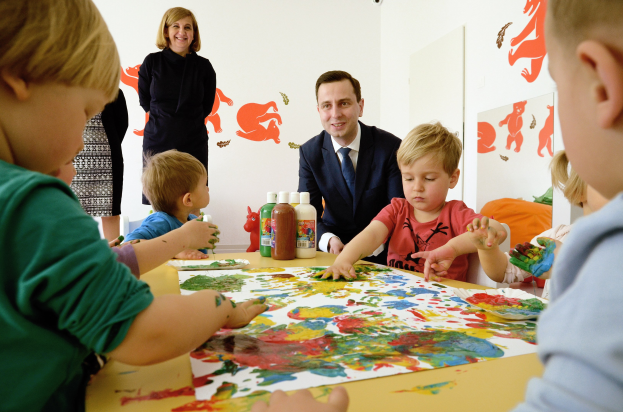 Gruppe von Kindern bei einer Malaktivität an einem Tisch mit bunten Händen, Papieren und Flaschen, beaufsichtigt von einer lächelnden Frau, mit Kunstwerken an der Wand im Hintergrund.