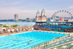 Großer Swimmingpool mit schwimmenden Menschen, umgeben von Geländern und Bänken, mit einem riesigen Riesenrad, Gebäuden, Bäumen und einem bewölkten Himmel im Hintergrund.