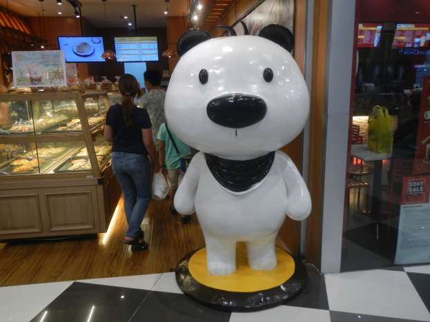 Eine Riesengänsepanda-Statue steht in einem Laden umgeben von Einkäufern, mit einer Auslage voller Lebensmittel links und Fernsehern an der Decke im Hintergrund.