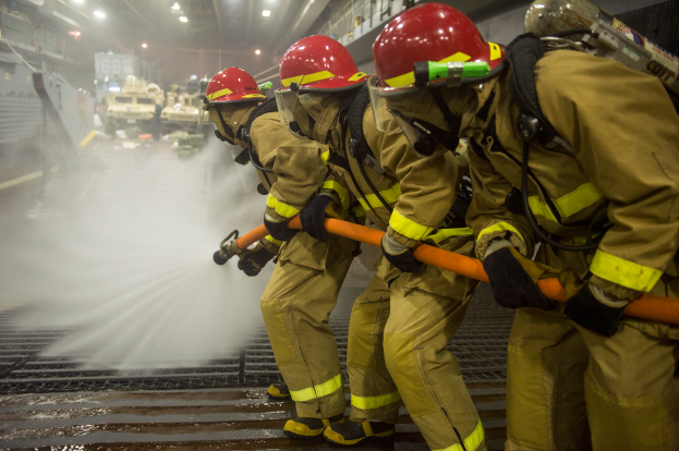 Feuerwehrleute in Helmen und Handschuhen halten Rohre und spritzen Wasser auf ein Feuerwehrauto, mit Lichtern, Geländern und Tafeln mit Text im Hintergrund.