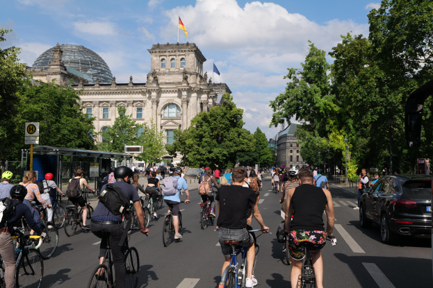 Eine Gruppe von Menschen, die auf Fahrrädern eine von Bäumen gesäumte Straße in Berlin, Deutschland, entlangfahren. Rechts befindet sich eine Bushaltestelle und auf einem nahen Gebäude weht eine Flagge.
