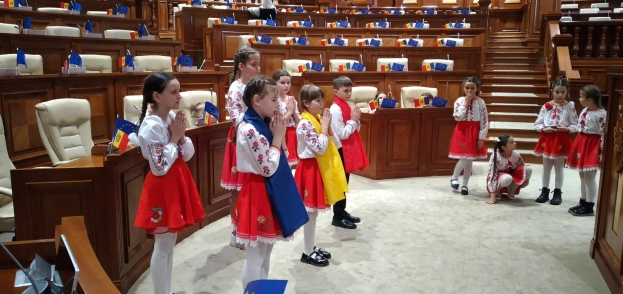 Eine Gruppe von Kindern hält Fahnen vor einem großen Raum mit Stühlen, Tischen und anderen Gegenständen, mit einigen Menschen, die sitzen und stehen, und einer Tür und einer Treppe auf der rechten Seite.