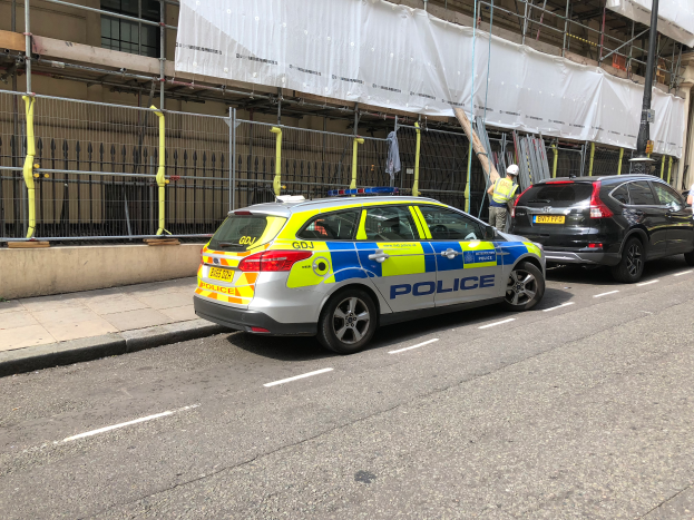 Ein Polizeiwagen steht an der Straßenseite neben einem Gebäude, zwei Helme tragende Personen stehen daneben; im Vordergrund sind ein Geländer und ein Pfahl zu sehen, und über das Gebäude ist ein weißes Tuch gehängt, was auf eine Polizeiuntersuchung nach einem Messerangriff in London hindeutet.