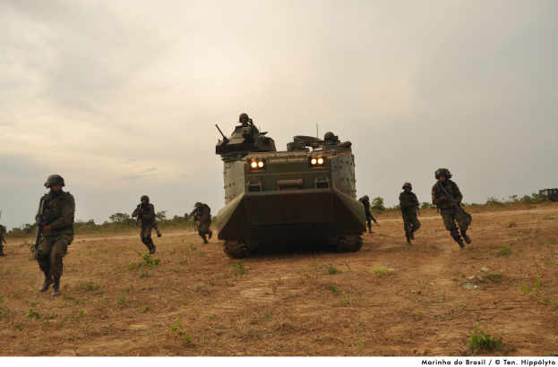 Eine Gruppe von Soldaten in Helmen und mit Gewehren marschiert über ein grasbewachsenes Feld neben einem gepanzerten Personentransporter, mit Bäumen und einem bewölkten Himmel im Hintergrund; Text ist unten im Bild sichtbar.