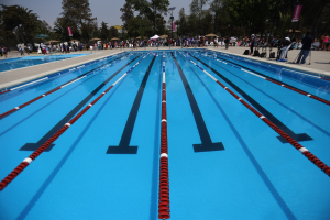 Großes Schwimmbad mit Bahnentrennern, umgeben von Menschen mit Taschen und Gegenständen, mit Zelten, Schildern, Stangen und einem bewölkten Himmel im Hintergrund.