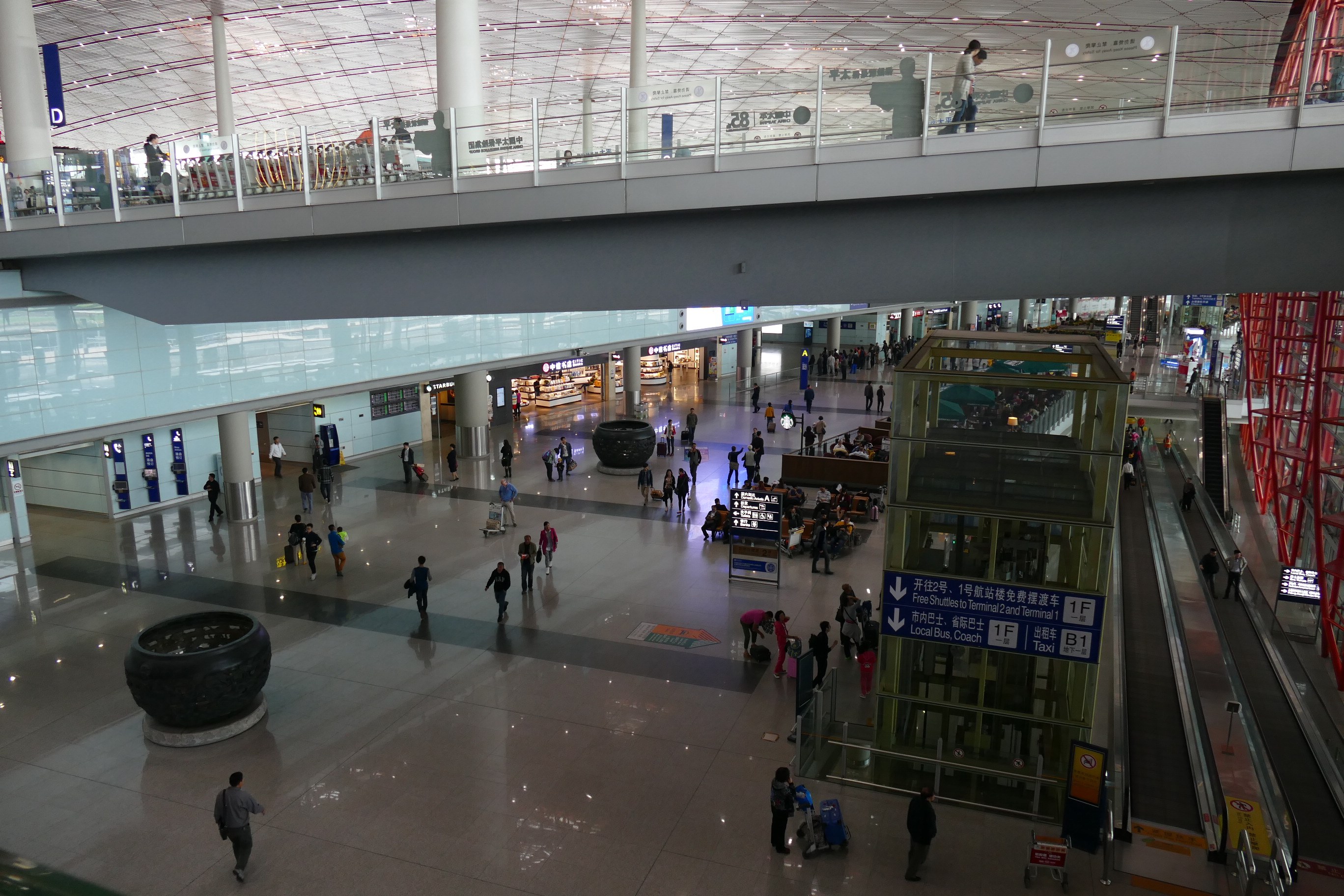 Eine große Gruppe von Menschen, die durch eine Flughafenhalle mit Rolltreppen, Informationsanzeigen, Säulen, Geländern und Deckenleuchten geht, wobei einige Taschen tragen.