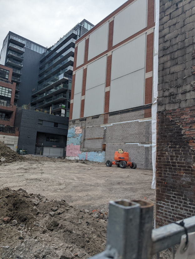 Baustelle mit einem teilweise abgerissenen Gebäude im Hintergrund, einem Fahrzeug, einem Metallzaun und einem bewölkten Himmel.