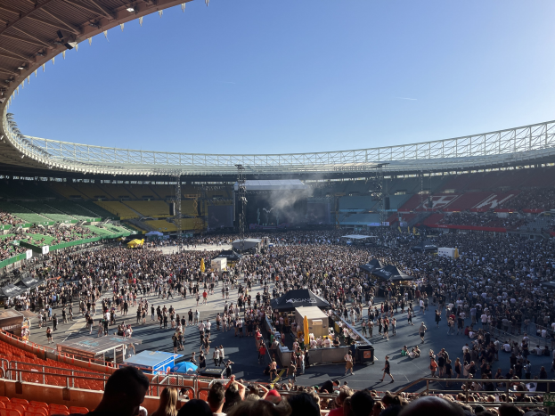 Große Menschenmenge in einem Stadion mit einer Bühne in der Mitte, mit Gegenständen und sichtbarem Himmel unter einem Dach mit Lichtern im Wembley-Stadion während eines Konzerts.