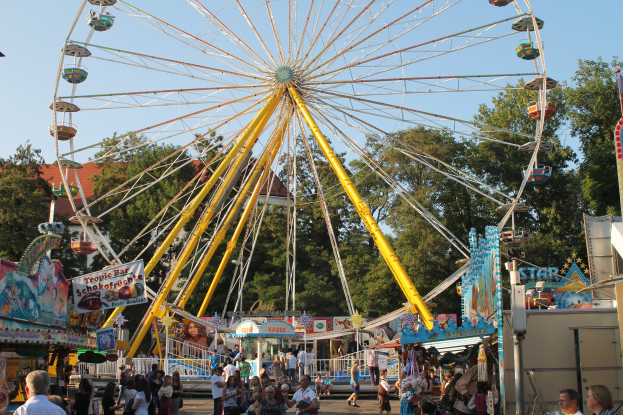 Ein großes Riesenrad auf einem Rummelplatz mit Menschen auf dem Boden, ein Stand mit Text auf der rechten Seite und Bäume mit einem klaren blauen Himmel im Hintergrund.
