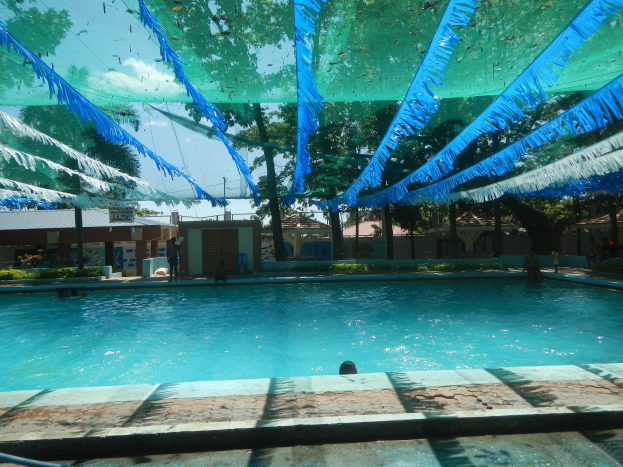 Ein großes Schwimmbad mit blauen und grünen Bändern an der Decke, umgeben von Häusern, Bäumen und einem klaren blauen Himmel, mit Menschen, die im Wasser und in der Sonne genießen.