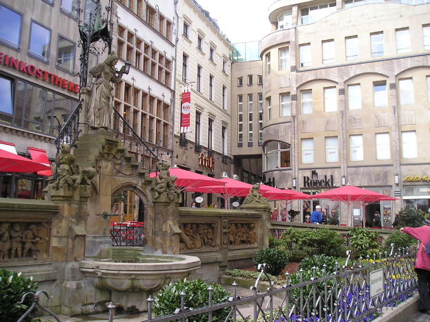 Eine Stadtansicht mit einem Teich, Pflanzen, einer Skulptur und einem Geländer im Vordergrund und Gebäuden, Zelten, Pflanzen und Menschen im Hintergrund.