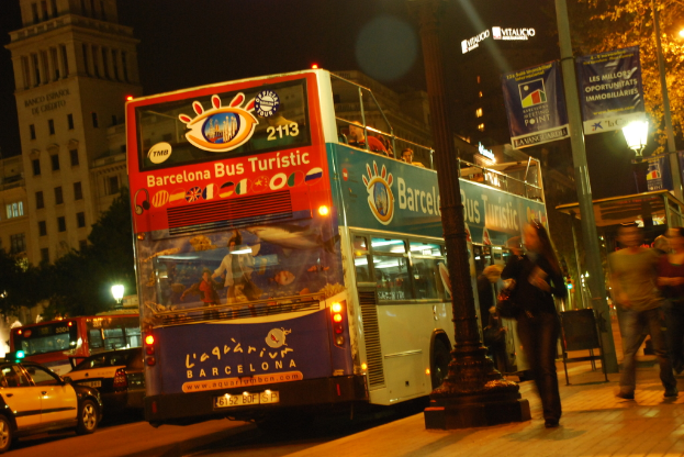 Fahrzeuge und ein Bus mit Fahrgästen fahren auf einer Straße, während Fußgänger auf dem benachbarten Gehweg gehen; Laternen, Banner, Bäume und Gebäude sind im Hintergrund zu sehen.