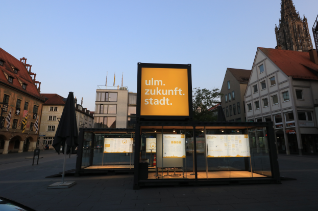 Eine Bushaltestelle auf einem Stadtplatz mit einem geparkten Auto davor, umgeben von Gebäuden, Bäumen und Fahnenmasten, mit einer Texttafel im Vordergrund und einem klaren blauen Himmel im Hintergrund.
