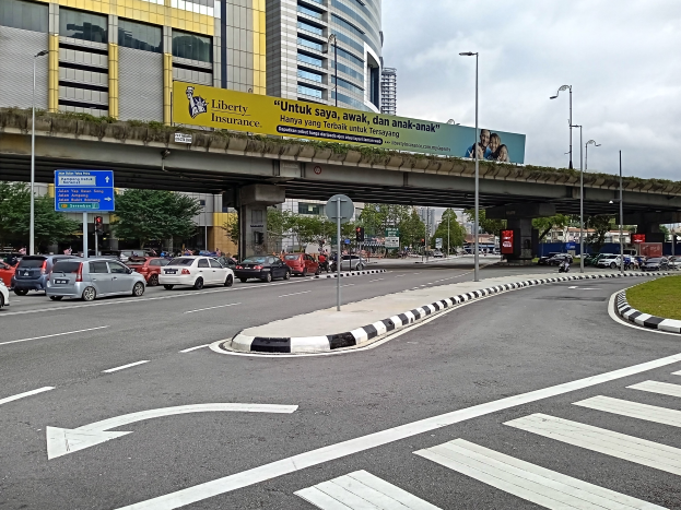 Eine Stadtstraßenszene mit Autos, einer Zebrastreifen, Gras, Laternen, einer Brücke mit Säulen, einer Werbetafel mit Text und Bildern, Bäumen, Gebäuden mit Fenstern und einem bewölkten Himmel.