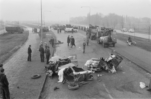 Schwarz-weiß-Szene eines Autounfalls am Straßenrand mit mehreren Fahrzeugen und einer Gruppe von Menschen drumherum, Laternenpfähle, Bäume und den Himmel im Hintergrund.