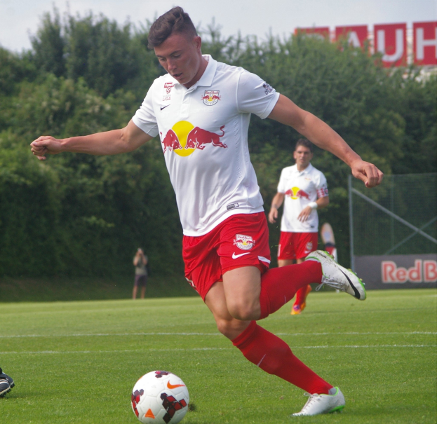 Ein Mann in einem Fußballtrikot schießt einen Ball auf einem grünen Feld mit Bäumen und einem klaren blauen Himmel im Hintergrund, wobei ein "RB Leipzig"-Schild sichtbar ist.