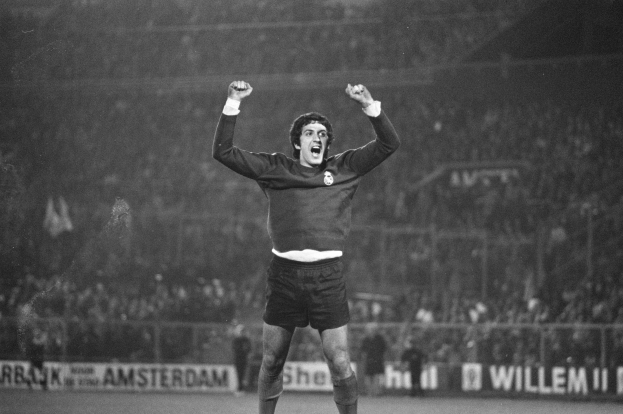 Ein Schwarz-Weiß-Foto eines Mannes, der auf einem Fußballfeld ein Tor feiert, mit erhobenen Armen und einem freudigen Gesichtsausdruck, umgeben von Menschen, einem Zaun und Bannern mit Text.