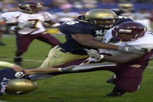 Zwei Fußballer mit Helmen und Uniformen kämpfen auf dem Feld gegeneinander, während Teamkollegen und Zuschauer im Hintergrund beobachten.
