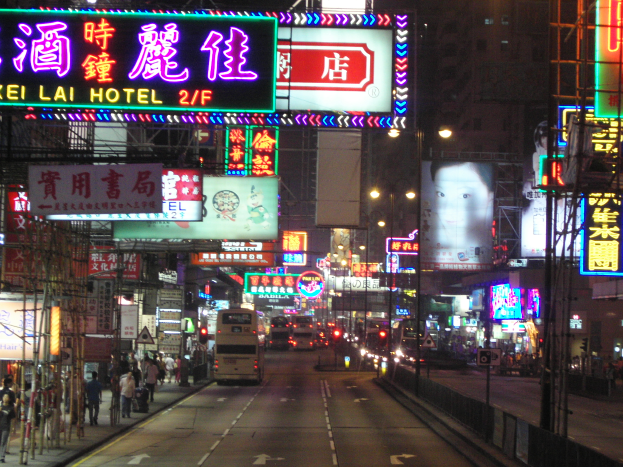 Eine nächtliche Stadtstraßenszene mit starkem Verkehr, Fußgängern, beleuchteten Gebäuden und verschiedenen Straßeneinrichtungsgegenständen.