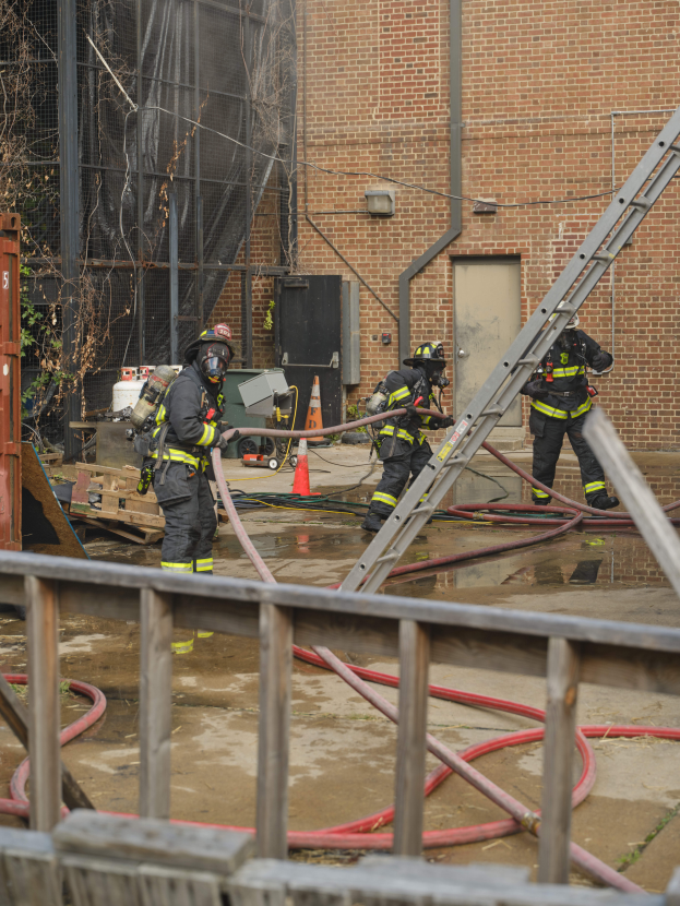 Eine Gruppe von Feuerwehrleuten in Helmen, die Rohre halten, arbeiten daran, ein Gebäude Feuer zu löschen, mit verschiedenen Ausrüstungen, Strukturen und einem Baum im Bild.