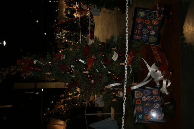 Ein Weihnachtsbaum mit Geschenken, Rahmen und Spielzeugen auf einer Plattform, mit Menschen, weiteren Weihnachtsb├Ąumen, Ketten, Masten und Lichtern im Hintergrund.