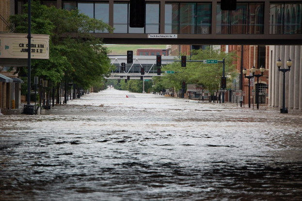 Überschwemmte Stadtstraße mit Wasser, das die Straße, Pfähle, Laternen, Schilder, Verkehrszeichen, Bäume, Gebäude und eine Brücke im Hintergrund bedeckt.
