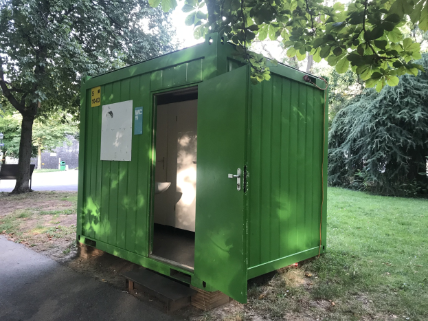 Grünes mobiles Klo mit einer Tür und einer angebrachten Platte, positioniert in einem Park umgeben von Bäumen und einer Bank auf der linken Seite, unter einem sichtbaren Himmel.