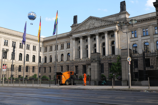 Das Reichstagsgebäude in Berlin, Deutschland, ein großes Gebäude mit zahlreichen Fenstern, umgeben von Straßenmasten, Fahrzeugen, Menschen und Bäumen, unter einem bewölkten Himmel.